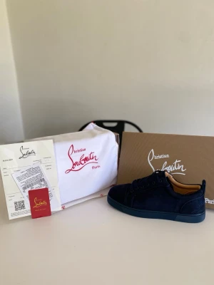 Mörkblå sneakers från Christian Louboutin - Snygga mörkblå sneakers i mocka från Christian Louboutin med ikonisk röd sula och ton-i-ton snörning. Skorna har en låg profil och klassisk siluett, perfekt för dig som vill sticka ut med exklusiv stil. Levereras med originalkartong och dustbag.