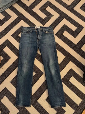 Blå raka jeans från Jeanerica,  - Väldigt öppen för förslag om pris.           Säljer ett par snygga blå jeans från Jeanerica i modellen CM002. Jeansen har klassisk femficksdesign, raka ben och knäppning med knappar. Tillverkade i 98% ekologisk bomull och 2% elastan för lite stretch. Perfekt blå tvätt med lätt slitning.