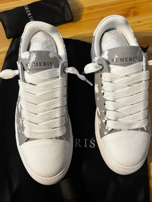 Numeris gråvita sneakers med mönster - Snygga sneakers från Numeris i grått och vitt med ett unikt kamouflagemönster. Skorna har tjocka vita snören, vit sula med räfflad undersida och tåparti i strukturmönstrat material. Perfekta för dig som vill sticka ut med stilrena detaljer.