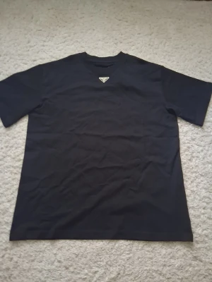 Svart t-shirt från Prada - Snygg svart t-shirt från Prada med det ikoniska metalltriangel-logotypen på bröstet. Klassisk rund halsringning och korta ärmar. Perfekt för dig som gillar stilrena designer och exklusiva märken.