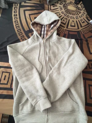 Grå zip hoodie Burberry - Säljer en grå zip hoodie från Burberry med klassiskt rutigt mönster i huvan. Hoodien har dragkedja framtill, två fickor och justerbar dragsko i huvan. Tillverkad i mjukt bomullsmaterial och har ribbade muddar vid ärmar och nederkant. XXL storlek men passar även för storlek M