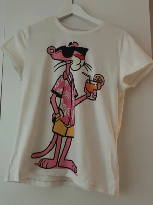 Vit Pink Panther t-shirt - Säljer en vit t-shirt med tryck av Pink Panther i semesterlook med solglasögon, drink och blommig skjorta. T-shirten har rund hals och korta ärmar. Perfekt för dig som gillar coola och lekfulla motiv.