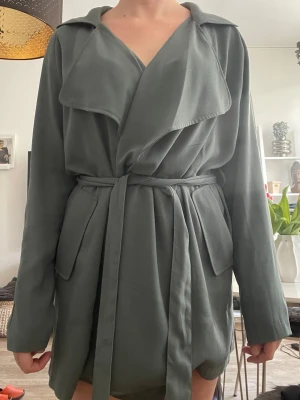 Tunn trenchcoat med skärp - Stilren grå/blå/grön trenchcoat med bred krage och knytskärp i midjan. Jackan har stora fickor framtill och ett mjukt, följsamt tyg som ger en avslappnad look. Perfekt för lager på lager och trendig till vardagsoutfits.
