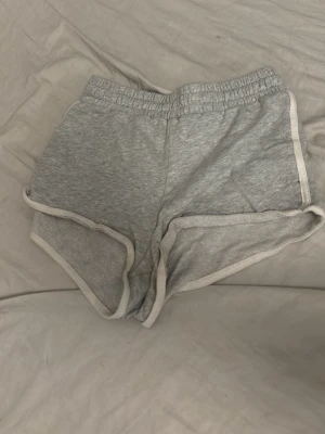 Grå sportiga shorts med vit kant - Grå shorts med elastisk midja och vita kanter. De har en sportig look och är tillverkade i mjukt bomullsmaterial, perfekta för avslappnade dagar eller träning.