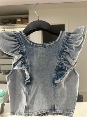 Blå volang topp i denim från Gina Tricot - Snygg blå denim topp från Gina Tricot med volangdetaljer längs axlarna och ärmlös design. Toppen har rund halsringning och dragkedja i ryggen för enkel på- och avtagning. Perfekt för dig som vill ha en trendig och unik look.