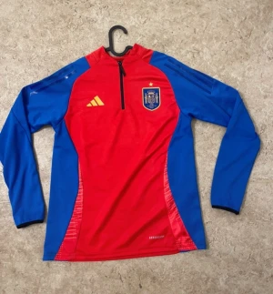 Spaniens träningströja Adidas röd/blå - Säljer en långärmad träningströja från Adidas i Spaniens landslagsfärger. Tröjan är röd med blå ärmar och sidor, har halv dragkedja och landslagets emblem på bröstet. Tillverkad i funktionsmaterial som andas. Perfekt för fotbollsträning.