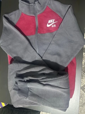 Nike Air mjukisbyxor grå och vinröd - Nike Air mjukisbyxor i grått med vinröda detaljer. Byxorna har resår och muddar vid benslut för en skön passform. Materialet är mjukt och bekvämt, perfekt för chill dagar. Matchande överdel med dragkedja och Nike-logga på bröstet.