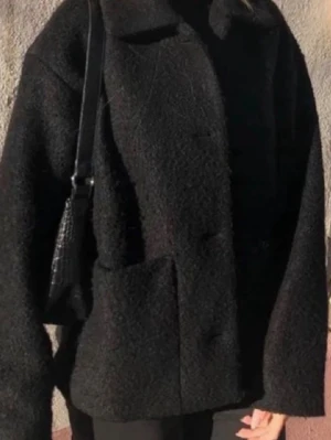 Svart kort kappa med krage - Säljer en kort svart kappa med oversized passform och klassisk krage. Jackan har stora fickor framtill och stängs med knappar. Perfekt för dig som gillar en stilren och mysig look. 