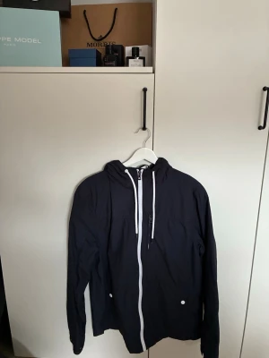 Marinblå vindjacka - Snyggmarinblå vindjacka med vit dragkedja och vita snören i huvan. Jackan har två fickor framtill med vita knappar och är tillverkad i ett lätt material som passar perfekt för blåsiga dagar. Enkel och stilren design som funkar till det mesta.