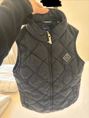 Mörk blå dunväst från GANT - Stilren mörk blå dunväst från GANT med quiltat mönster och hög krage. Västen har dragkedja framtill och en diskret logotyp. Bra och enkel att matcha med olika outfits.