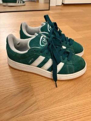 Adidas Campus gröna sneakers - Gröna Adidas Campus sneakers i mocka med klassiska vita ränder på sidorna och vit sula. Skorna har gröna skosnören och en vit Adidas-logga på plösen. Insidan är fodrad i ljusgrått tyg. Perfekta för dig som gillar retrostil och streetwear. De är sparsamt använd men det finns en skavank inuti ena skon (se bild) annars superfint skick och pris kan absolut diskuteras ❤️