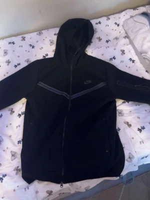 Svart Nike tech hoodie med dragkedja - Snygg svart hoodie från Nike med hel dragkedja framtill och huva. Tröjan har diskreta reflexdetaljer över bröstet och en liten Nike-logga på vänster sida. Perfekt för dig som gillar sportig och stilren streetwear.