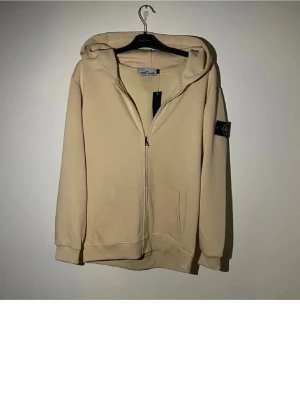 Beige hoodie från Stone Island - Snygg beige hoodie från Stone Island med dragkedja framtill och klassisk logopatch på vänster ärm. Hoodien har huva, två fickor fram och ribbade muddar. Perfekt för en avslappnad streetwear-look.