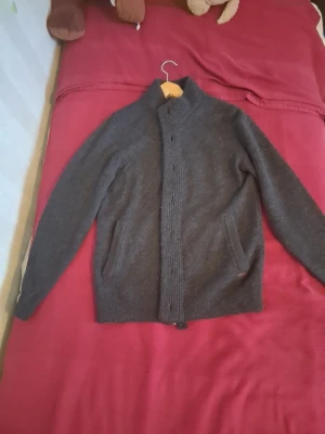 Mörkgrå kofta från Barbour - Mysig mörkgrå kofta från Barbour med hög krage och dragkedja framtill. Koftan har två fickor på sidorna och är stickad i ett mjukt material. Perfekt för lager på lager och en stilren look.