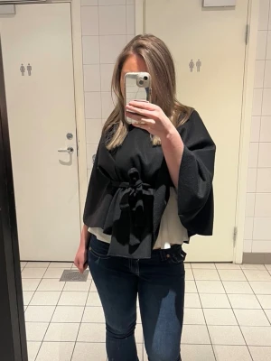 Svart kappa med knytband - Stilren svart kappa från Missay med knytband i midjan och en knapp vid halsen. Kappan har vida ärmar och en mjuk, filtad yta som ger en elegant look. Perfekt för lager-på-lager och enkel att matcha med olika outfits.