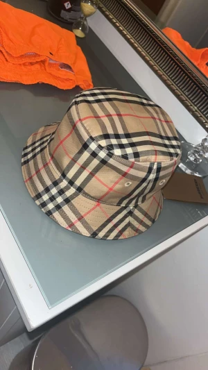 Burberry rutig bucket hat beige - Priset kan diskuteras vid dm.                  Säljer en snygg bucket hat från Burberry med klassiskt rutigt mönster i beige, svart, vitt och röda linjer. Hatten har en mjuk form och är tillverkad i bomull med svart innertyg. Perfekt för att lyfta din streetwear-look. Size M passar S och L. UTAN KVITTO