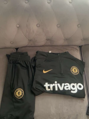 Chelsea FC svart träningsset Nike - Säljer ett svart Chelsea FC träningsset från Nike med guldfärgade detaljer. Setet består av en långärmad tröja med halv dragkedja och byxor, båda med Chelsea-logga och Nike swoosh. Materialet är mjukt och stretchigt, perfekt för träning eller chill.