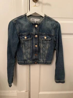 Cropped jeansjacka från Zara - Snygg croppad jeansjacka från Zara i tvättad blå denim. Jackan har rund halsringning, guldiga knappar framtill och två bröstfickor med lock. Klassisk modell med raka ärmar och markerad midja. Perfekt för en trendig och avslappnad look. Använd endast 1 gång. 
