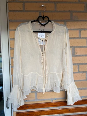 Krämvit volangblus Moss Copenhagen - Luftig krämvit blus från Moss Copenhagen i transparent chiffong. Blusen har långa ärmar med volangdetaljer, knytning framtill och flera lager volanger nedtill och baktill. Perfekt för en romantisk och bohemisk stil.