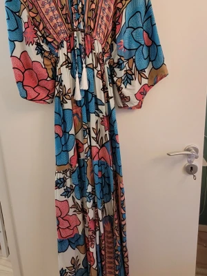 Blommig långklänning med snörning från Mexiko i Nyskick. - Färgstark sommarklänning med stora turkosa och rosa blommor på vit botten. Klänningen har snörning med tofsar  och vida ärmar med fickor i sidorna. Perfekt för dig som gillar bohemiska och uttrycksfulla plagg. Dyr i inköpspris. Använd 2 ggr. 100% Bomull 