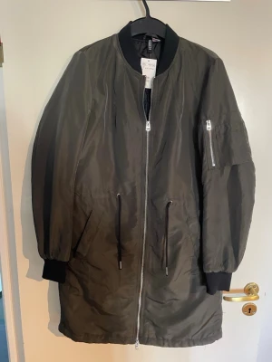 Lång grön vindjacka från H&M - Stilren lång vindjacka i mörkgrönt från H&M. Jackan har dragkedja framtill, svarta muddar vid ärmslut och hals, samt snörning i midjan. Praktiska fickor och en ärmficka med dragkedja. Perfekt för blåsiga dagar och enkel att matcha.