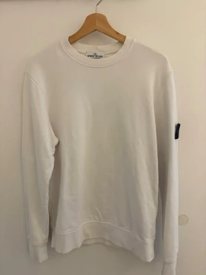 Vit sweatshirt från Stone Island - Vit sweatshirt från Stone Island med klassisk rund halsringning och ribbade muddar. Tröjan har den ikoniska svarta logotypen på vänster ärm. Perfekt för en clean och stilren look.