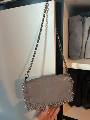 Grå axelremsväska från Tiamo - Snygg grå handväska från Tiamo med kedjerem och dekorativa metallkedjor runt kanterna. Väskan är i konstläder och har ett fack med dragkedja på insidan. Perfekt storlek