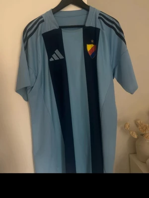 Djurgården matchtröja Adidas blå - Säljer en matchtröja pga att har en dublett av exakt samma tröja🤗Snygg ljusblå Djurgården matchtröja från Adidas med mörkblå ränder och klubbmärke på bröstet. Tillverkad i lätt och ventilerande polyester, perfekt för träning eller match. Klassisk sportig design med korta ärmar och rund hals.