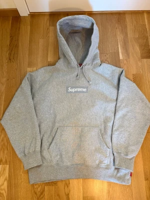 Grå Supreme hoodie med box logo FW24 - Säljer en grå hoodie från Supreme med klassisk box logo broderad på bröstet. Hoodien har stor huva med dragsko, magficka och ribbade muddar. Tillverkad i mjuk bomullsblandning som är perfekt för chill dagar. Ända skavanker är slitage på ena armmudden (kolla bild) men inget utöver det. I storlek M.