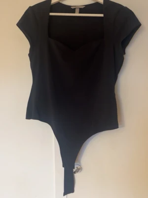Svart body med fyrkantig urringning - Snygg svart body med korta ärmar och fyrkantig urringning. Modellen har en markerad midja och klassisk knäppning nedtill. Perfekt att styla med jeans eller kjol för en trendig look. Den är i S men känns större så skulle säga M