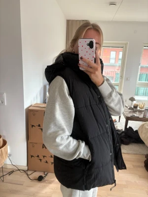 Svart dunväst med huva - Snygg svart dunväst med huva och tryckknappar framtill. Västen har en puffig, oversized passform och är perfekt att slänga över en hoodie. Inga synliga loggor eller märken. Storlek M