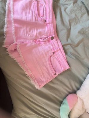 Ljusrosa fransade jeansshorts - Säljer ett par ljusrosa jeansshorts med fransade kanter och klassisk femficksdesign. Shortsen har knapp och dragkedja framtill och är perfekta för varma dagar. Supersnygga till sneakers eller sandaler! Köpte de nyss säljer för att de är fel storlek 