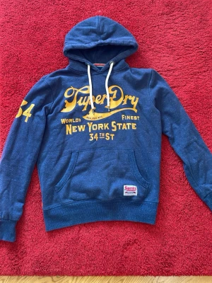 Blå Superdry hoodie med tryck - Snygg blå hoodie från Superdry med gul text och tryck på bröstet och ärmen. Storlek M.