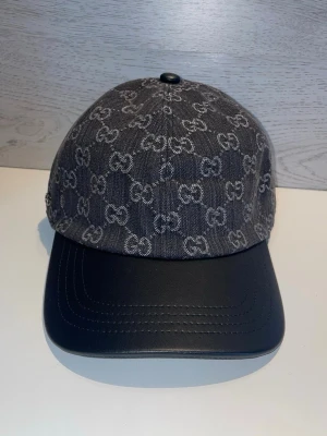 Gucci keps i denim och läder - Snygg keps från Gucci med mörkgrå denim och klassiskt GG-mönster i vitt. Skärmen är i svart läder för en exklusiv känsla. Perfekt accessoar för dig som vill sticka ut med stilrena detaljer och lyxig vibe.