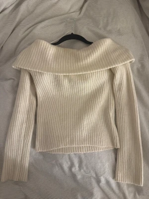 Off shoulder stickad tröja cremevit - Trendig cremevit stickad tröja med bred off shoulder-krage och ribbad struktur. Långa ärmar och figurnära passform ger en snygg siluett. Perfekt för dig som vill ha en stilren och modern look till hösten eller vintern.
