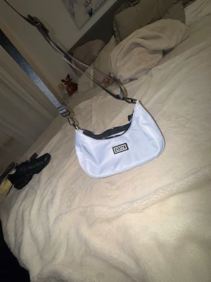 Ljusblå axelväska från CMMN SWDN - Snygg ljusblå axelväska från CMMN SWDN med svart justerbar axelrem och dragkedja. Väskan har en stilren form, svart innerfoder och en praktisk innerficka. Perfekt för att bära det viktigaste och ger en trendig touch till din outfit.