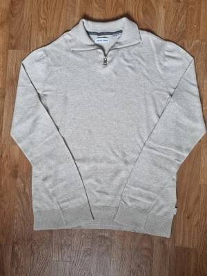 Beige halvzip tröja Jack & Jones - Snygg beige tröja från Jack & Jones med halv dragkedja och ståkrage. Tillverkad i mjukt material, perfekt för lager på lager. Långärmad modell med ribbade muddar vid ärmslut och nederkant. Enkel och stilren design som passar till det mesta.