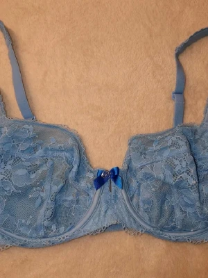 Blå spets-bh med rosett - Söt blå bh i spets med blommigt mönster och justerbara axelband. Dekorerad med en liten blå rosett och en glittrig detalj framtill. Bh:n har bygel och hakknäppning bak för extra stöd och passform. Perfekt för dig som gillar romantiska detaljer.