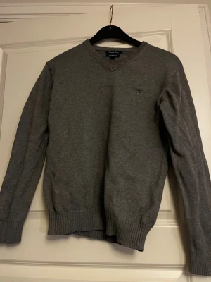Grå stickad tröja Gant stl 158/164 - Klassisk grå stickad tröja från Gant med V-ringning och diskret logga på bröstet. Tröjan har långa ärmar och ribbade muddar vid ärmslut och nederkant. Perfekt för lager-på-lager och enkel att matcha. Stl 158/164