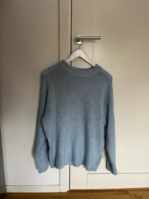Ljusblå stickad tröja - Mysig ljusblå stickad tröja med rund halsringning och långa ärmar. Tröjan har en enkel, rak passform och ribbstickade muddar vid ärmslut och nederkant. Perfekt för lager på lager under kyliga dagar.