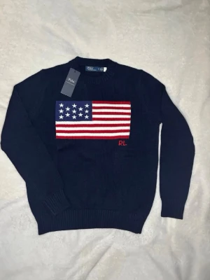 Mörkblå stickad tröja Polo Ralph Lauren - Snygg mörkblå stickad tröja från Polo Ralph Lauren med stor amerikansk flagga i rött, vitt och blått på bröstet samt RL-broderi. Tröjan har rund halsringning och ribbade muddar. Perfekt för dig som vill ha en klassisk och trendig look.