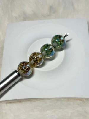 Färgglada glaspärlor till Penna 16mm - Fyra runda glaspärlor i gröna och guldiga toner med detaljerade mönster och metallinslag. Pärlorna har en skimrande yta med guldkrona inuti. 16 mm och passar penna 