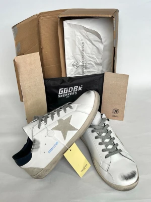 Golden Goose vita sneakers med stjärna - Golden Goose sneakers i vitt läder med grå mockadetaljer, silverfärgad stjärna på sidan och grå snören. Skorna har en vintage-look med slitningar på tån och sulan samt svart hälparti. Klassisk låg modell med GGDB-logga på sidan.