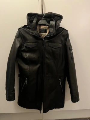 Svart skinnjacka med huva KappAhl - Snygg svart skinnjacka från Goldstein , fyra framfickor med dragkedja och lock samt en extra ficka på ärmen. Lång modell med raka linjer och coola detaljer i metall. Perfekt för dig som vill ha en stilren och edgy look. Står 42 i jackan men den är liten i storlek passar M/L