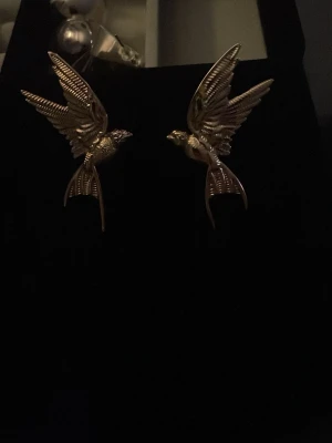 Guldiga örhängen i form av svalor - Unika örhängen från ur & Penn med detaljerad design av svalor i flykt. De är guldiga och har en cool, retrovibe som verkligen sticker ut. Perfekta för dig som vill ha något annorlunda och snyggt till din stil.
