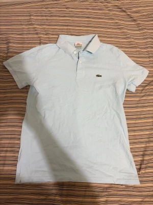 Lacoste pike polotröja - Klassisk ljusblå pikétröja från Lacoste med korta ärmar och krage. Tröjan har två knappar framtill och den ikoniska krokodilloggan broderad på bröstet. Tillverkad i bomull för en bekväm och luftig känsla.