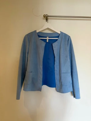 Ljusblå kavaj från H&M - Stilren ljusblå kavaj från H&M i mjukt mockaimitationsmaterial. Kavajen har öppen front utan knäppning, två stora fickor fram och rundad halsringning. Perfekt för en trendig och avslappnad look.