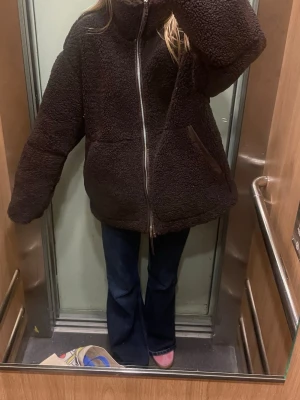 Mörkbrun oversized teddyjacka - Mysig och oversized teddyjacka i mörkbrunt med hög krage och grov silverfärgad dragkedja. Jackan har två snedställda fickor och ett fluffigt, fårskinnsinspirerat material som ger en riktigt chill vibe. Perfekt för kalla dagar när du vill vara både varm och snygg.