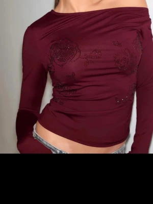 Vinröd offshoulder topp med blommönster - Trendig vinröd offshoulder topp med långa ärmar och blommönster i form av små glittriga stenar. Sitter tight och har en croppad passform som framhäver midjan. Perfekt för dig som vill sticka ut med en unik detalj på din outfit.