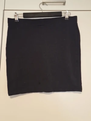 Svart enkel kortkjol i bomull - En svart, enkel och rak kortkjol i mjukt bomullsmaterial. Kjolen har en klassisk och stilren design utan synliga detaljer eller mönster, vilket gör den lätt att matcha med olika toppar och accessoarer.